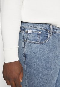 Vaqueros de mezclilla azul claro con un diseño clásico de cinco bolsillos, con un logo cK en el bolsillo trasero y un acabado de tela texturizada.