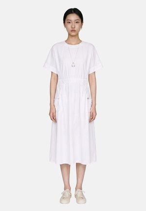 Femme debout, de face, portant une robe midi blanche à manches courtes, des baskets beiges et un collier avec un pendentif.