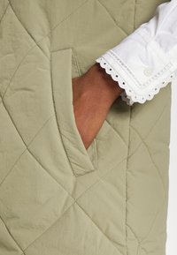 Gekwijlde olijfgroene kledingstukken met een zakdetail, met een witte oogjesblouse die aan de manchette tevoorschijn komt. Soepele textuur.