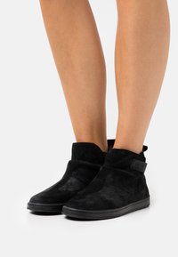 Cizme negre din suede la glezna, cu o textură moale, având un design cu pliuri și un patch logo negru pe lateral. Talpă din cauciuc plat.