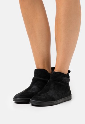 Bottines noires en daim avec une texture douce, présentant un design à rabat et un patch logo noir sur le côté. Semelle en caoutchouc plate.