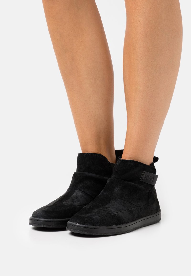 Cizme negre din suede la glezna, cu o textură moale, având un design cu pliuri și un patch logo negru pe lateral. Talpă din cauciuc plat.
