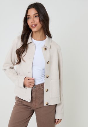 Kobieta o długich ciemnych włosach, ubrana w beżową kurtkę zapinaną na guziki, białą bluzkę i brązowe spodnie, stoi na tle jednolitego jasnego tła.