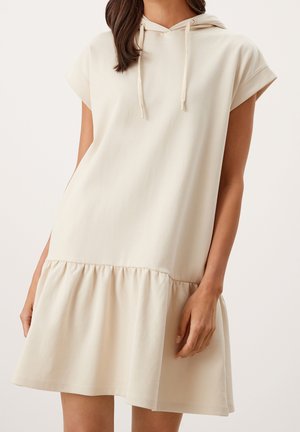 Kvinde iført en kortærmet, beige hættetrøje-kjole med en samlet nederdel og snor ved halsudskæringen, stående mod en ensfarvet baggrund.