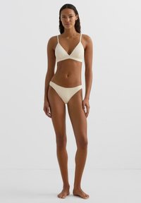 Ženska stoji bosa v teksturiranem smetanasto belem bikiniju z V-izrezom na zgornjem delu in ujemajočih visokim krojem hlačicah na enostavni belem ozadju.