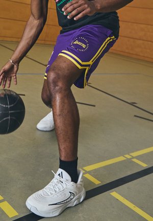 Person iført lilla Lakers-shorts, der dribler en sort basketball på en indendørs bane med hvide sneakers og sorte sokker.