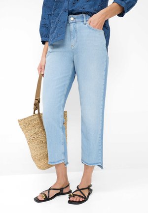 Kvinde iført lyseblå cropped jeans, blå tekstureret bluse, sorte sandaler med stropper og gulddetaljer, og holder en flettet stråtaske.