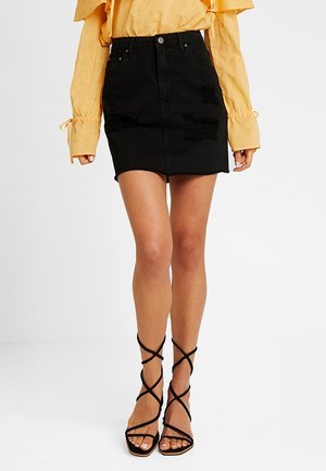 Vrouw die een zwarte versleten denim mini-rok draagt, een gele blouse met lange mouwen en zwarte, met bandjes versierde hoge hakken, staand tegen een witte achtergrond.