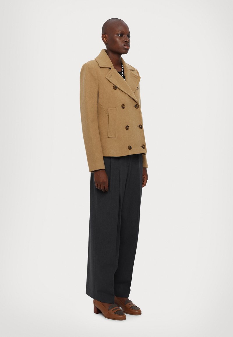 Kamel-farbenen, doppelt geknöpften Wollmantel mit großen braunen Knöpfen, seitlichen Taschen und einem Reverskragen, kombiniert mit grauen, weit geschnittenen Hosen.