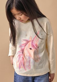 Chemise beige à manches longues avec un graphique de licorne, crinière rose et accents scintillants, associée à un jean bleu. Texture lisse, coupe décontractée.