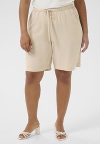Beige relaxed-fit shorts with an adjustable drawstring waistband, side pockets, and smooth texture, styled with white block-heeled sandals.