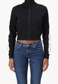 Schwarze, kurz geschnittene Jacke mit Reißverschluss und hohem Kragen, mit weißen Akzenten entlang der Ärmel; kombiniert mit blauen Jeans. Glatte Stoffstruktur.