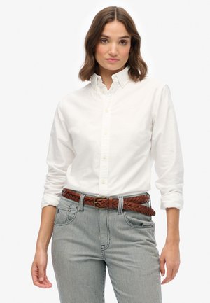 Una mujer con cabello castaño ondulado lleva una camisa blanca abotonada, pantalones a rayas y un cinturón trenzado marrón, todo contra un fondo blanco liso.