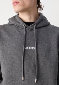 Vue partielle d'une personne portant un sweat à capuche gris avec « LES DEUX » imprimé en blanc sur la poitrine, par-dessus une chemise noire.