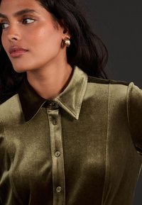 Oliwkowa, welurowa koszula z wysmukłym kołnierzem, z zapięciem na guziki i wyraźną fakturą. Uzupełniona złotymi kolczykami w kształcie kół.