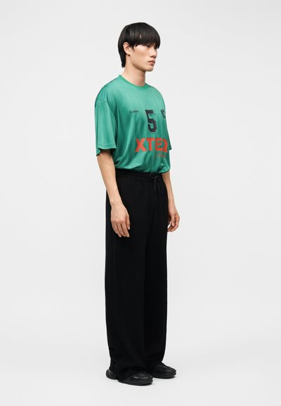 Νέος άνδρας που στέκεται πλάγια, φοράει φαρδιά πράσινη μπλούζα με αριθμό 5 και μαύρο παντελόνι φαρδιάς γραμμής, με λευκό φόντο.