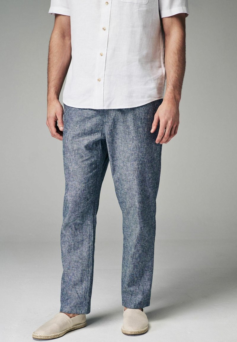Homme portant une chemise blanche à manches courtes boutonnée, un pantalon en lin bleu et des chaussures beiges à enfiler, debout devant un fond uni.
