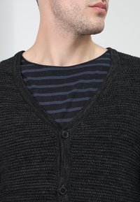 Svart texturerad kofta med V-ringning, försedd med två knappar. Bärs över en marinblå och svart randig t-shirt.