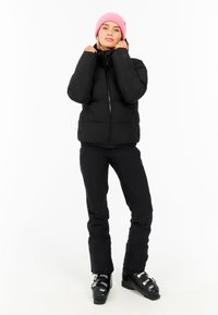 Zwarte pufferjack met een hoge kraag, voorzien van een ritssluiting en zijzakken. In combinatie met zwarte broek en een roze gebreide muts. Zwarte skischoenen.