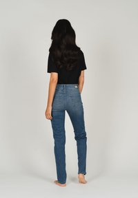 Jeans in denim blu con una vestibilità slim, vita alta e due tasche posteriori. Indossati con una t-shirt nera, a piedi nudi, su uno sfondo bianco.