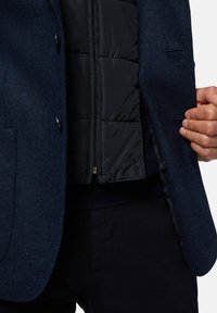 Blazer in tessuto a spina di pesce blu navy con gilet imbottito nero all'interno. Presenta due bottoni, una tasca interna e un esterno in lana lavorata.