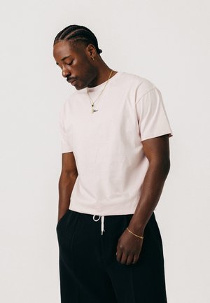 Mann mit geflochtenem Haar, der ein hellrosa T-Shirt, schwarze Hose mit weißem Kordelzug, goldene Halskette, Armband und Ohrring trägt und nach unten schaut.
