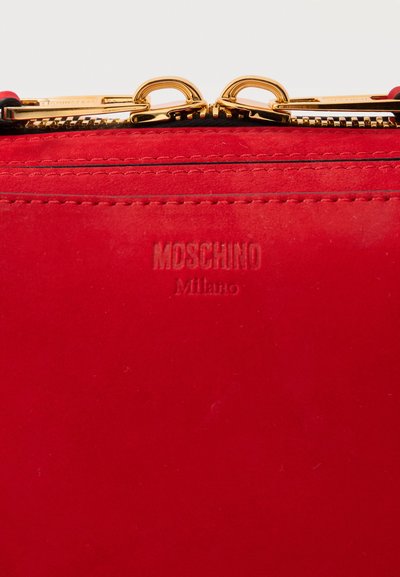 Κόκκινο δερμάτινο αντικείμενο Moschino Milano με ορατές ραφές και δύο χρυσά φερμουάρ, μερικώς κλειστά στο κέντρο της κορυφής.