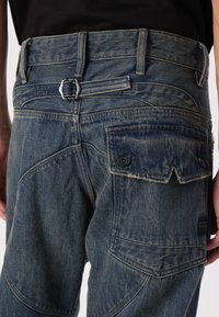 Jeans en denim bleu avec une taille ajustée, un détail de boucle en argent, des coutures contrastées, des poches cargo et une finition texturée.