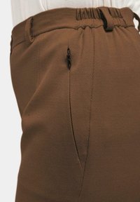 Braune Hose aus strukturiertem Stoff mit einem elastischen Bund und einer seitlichen Reißverschlusstasche. Sichtbare Detailnähte entlang der Nähte.
