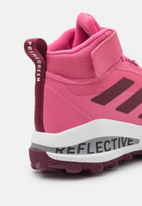 Różowy but sportowy o wysokiej cholewce, z teksturowanym materiałem górnym, odblaskowymi akcentami oraz wyraźnymi czarnymi paskami. Wyposażony w wyściełany kołnierz i pętelkę na pięcie.
