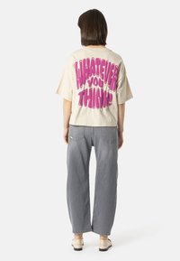 Osoba s krátkými hnědými vlasy stojí zády, má na sobě béžové tričko s růžovým nápisem "CO MYSLÍTE!" a šedé široké džíny.