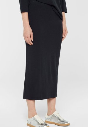 Frau trägt einen schwarzen Midirock kombiniert mit silbernen und weißen Sneakern, steht vor einem einfarbigen hellen Hintergrund.