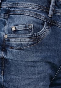 Denimjeans med en blå tvätt, med en bakficka, silvernitar och synlig sömnad. Slät yta med lätt blekning.