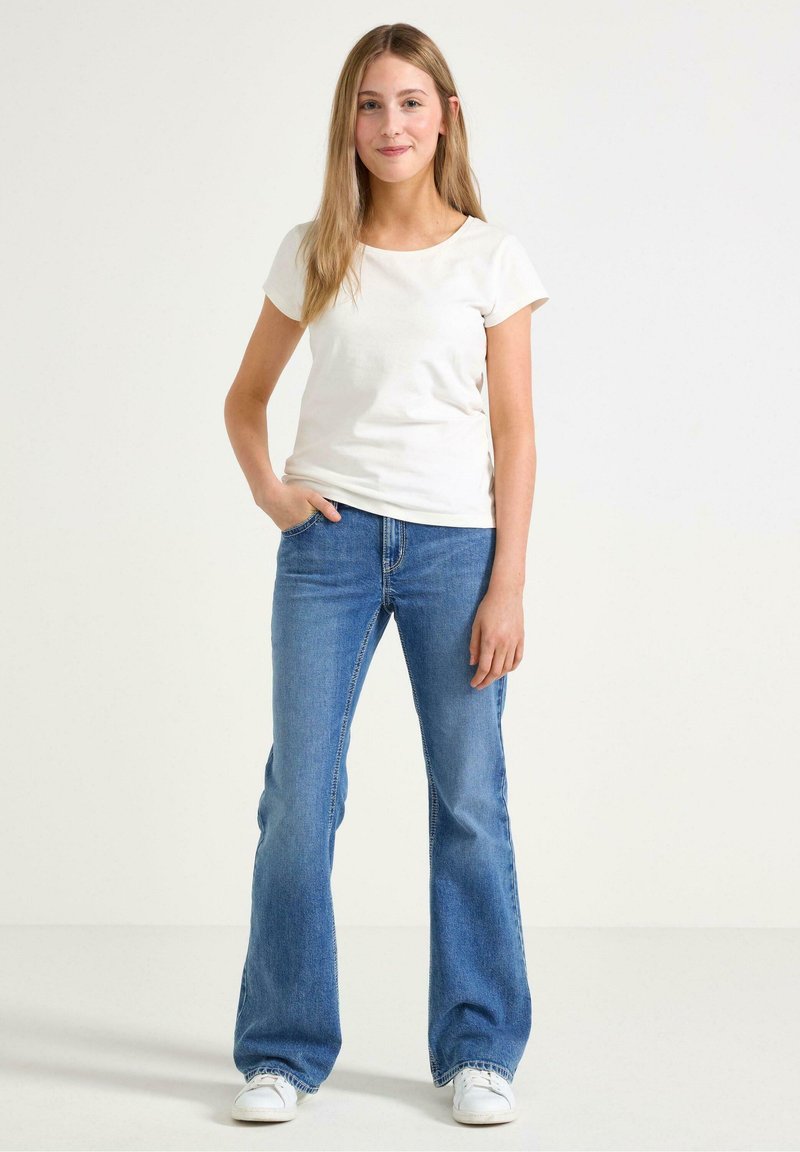 Vit t-shirt med kort ärm i kombination med blå utsvängda jeans. Jeansen har en klassisk passform med sömdetaljer och bärs med vita skor.