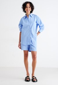 Lyseblå, knappet skjorte med rullede ermer, kombinert med matchende shorts. Antrekket har en glatt tekstur og avslappet design. Sorte sandaler fullfører looken.