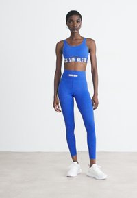 Femme portant un brassière de sport bleu Calvin Klein et un legging taille haute avec des baskets blanches, se tenant devant un mur blanc uni.