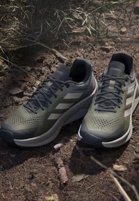 Gröna träningsskor med ett meshnät ovan med perforeringar, grå detaljer och tre vita ränder. Utrustade med en vadderad sula.