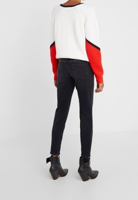 Persona vestida con pantalones vaqueros negros, botines negros y un suéter de punto blanco con acentos rojos y negros en las mangas, de pie contra un fondo simple.