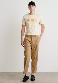 T-shirt crème avec un texte audacieux "HUGO" en jaune, associé à un pantalon beige. Une casquette noire et des mocassins noirs complètent la tenue.
