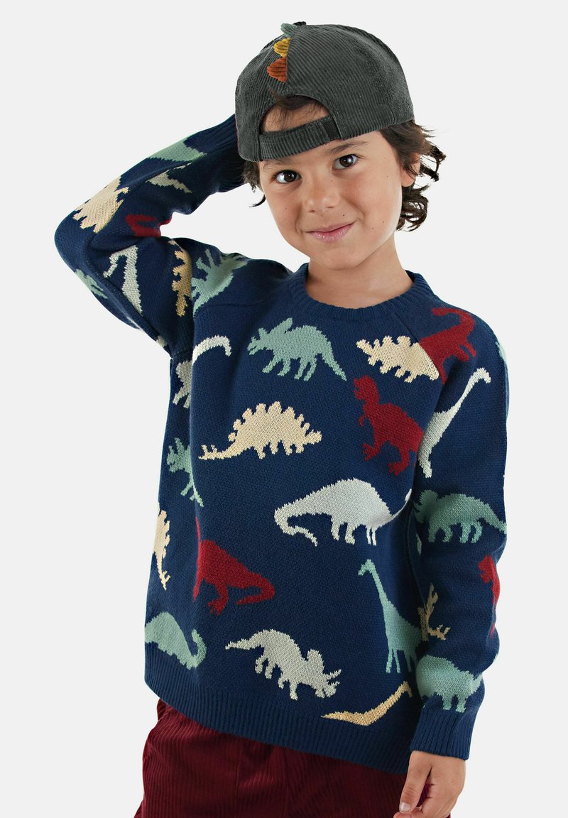 Junge trägt eine dunkelgrüne Kappe rückwärts, einen marineblauen Pullover mit bunten Dinosauriermustern und rote Hosen und posiert mit der Hand auf dem Kopf.