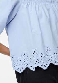 Lys blå bomullsbluse med løs passform, rynket halslinning og bølget broderi langs kanten.