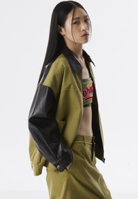 Veste vert olive avec des manches en cuir noir, design oversize, poches frontales, assortie à un top court avec des broderies rouges.
