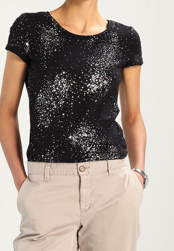 Personne portant une chemise noire à manches courtes avec des motifs d’étoiles blanches et un pantalon beige, mains dans les poches, sur un fond clair uni.