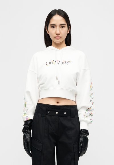 Κοντοκομμένο λευκό φούτερ με κουκούλα και λογότυπο με λουλούδια στο μπροστινό μέρος, μακριά μανίκια με πολύχρωμα λουλουδένια σχέδια, σε συνδυασμό με μαύρο παντελόνι και γάντια.