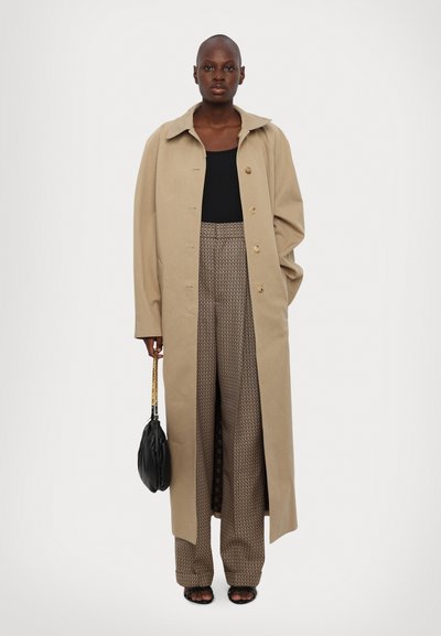 Bež dolg plašč z velikim ovratnikom in sprednjimi gumbi, v kombinaciji z vzorčastimi širokimi hlačami. Model drži črno torbico in nosi sandale.