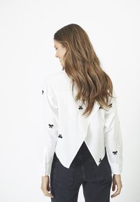 Camicia bianca con bottoni decorata con ricami a forma di fiocco neri, progettata con un originale taglio sulla schiena e una vestibilità leggermente oversize. Abbinata a jeans neri.