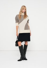 Gruba dzianinowa sweter w kremowo-taupe paski, zestawiony z czarną spódnicą do kolan i czarnymi kozakami za kolano, trzymając czarną torebkę.