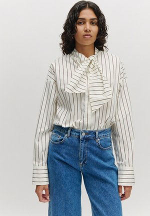 Weiße gestreifte Bluse mit einem Schleifenkragen, langen Ärmeln und Knopfverschluss, kombiniert mit hochgeschnittenen blauen Jeans.