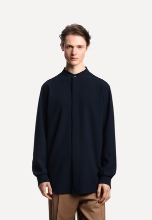 Giovane uomo con pelle chiara e capelli castani indossa una camicia button-down di un blu navy scuro e pantaloni color sabbia, in piedi contro uno sfondo bianco semplice.