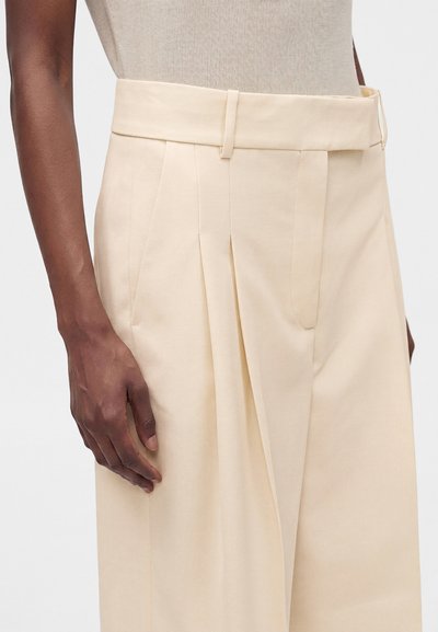 Gros plan d'une personne à la peau foncée portant un pantalon beige à jambes larges avec plis et taille haute, associé à un haut sans manches beige clair.